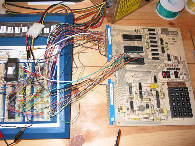 A KIM-1 next to a breadboard with some circuitry on it.  There are many wires going from the KIM-1 to the breadboard, some from the usual side connectors, but also some directly from one of the 40-pin IC sockets on the KIM-1.  The KIM-1 is missing 3 seven-segment display, but the leftmost one is on and shows a 0
