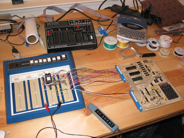 A KIM-1 on a workbench connected to a breadboard with a several wires.  The breadboard has some circuitry with a ZIF socket holding an EPROM. There are many EPROMs with various labels set aside on the breadboard unit.