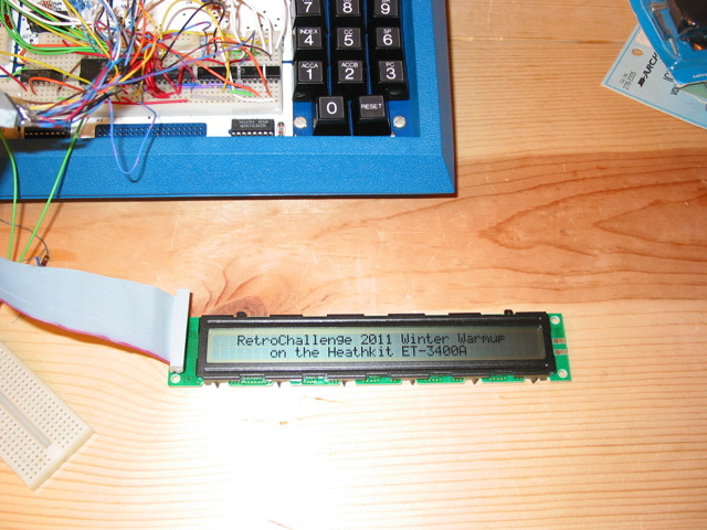 The bottom part of a Heathkit ET-3400A Microprocessor trainer which has some circuitry on it, connected to a two-line LCD display module.  The module shows: RetroChallenge 2011 Winter Warmup on the Heathkit ET-3400A