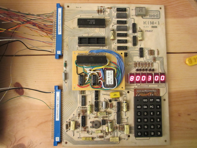 A KIM-1 with some wires heading away from the connectors.  There is a homebrew daughter board hovering above one of sockets where there would be a RRIOT.  The other RRIOT socket is empty.  The seven-segment displays are bright and show 8003 00.  Two of the memory chips are not like the others and are in sockets.